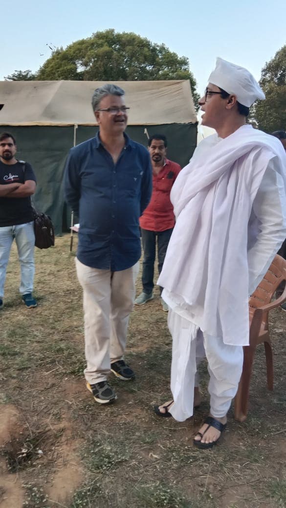 Two men in conversation beside a crew tent, one in white khadi costume